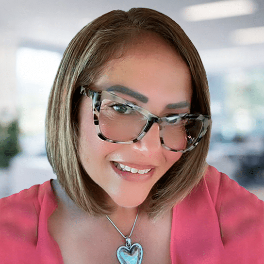 A woman with shoulder-length brown hair, wearing large tortoiseshell glasses, a pink top, and a silver heart-shaped pendant, smiles in a brightly lit, blurred office setting.