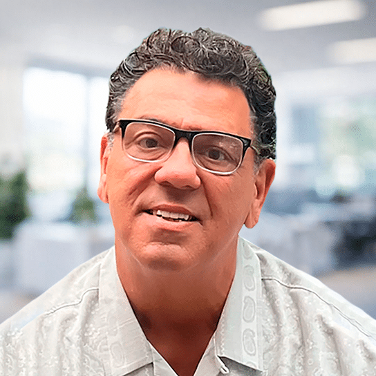 A smiling man with short curly hair and glasses wearing a light-colored, patterned shirt, standing in a bright office setting with blurred desks and windows in the background.