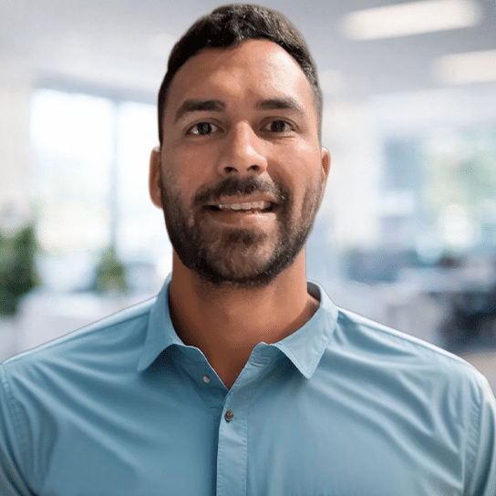 A man with short dark hair and a beard, wearing a light blue collared shirt, smiles in a bright office setting with large windows and blurred background.