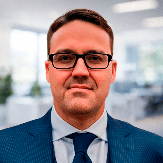 A man wearing glasses and a blue suit with a tie is standing in an office setting, looking directly at the camera. The background is blurred, showing windows and office furniture.