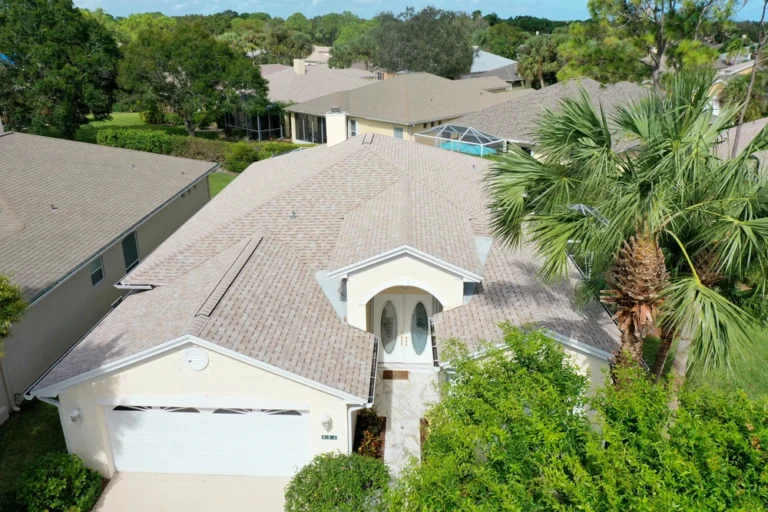 Aerial view of a light-colored house with a two-car garage, surrounded by trees and greenery. The roof is sloped, and the entrance features a small porch area. Other similar houses are visible nearby.