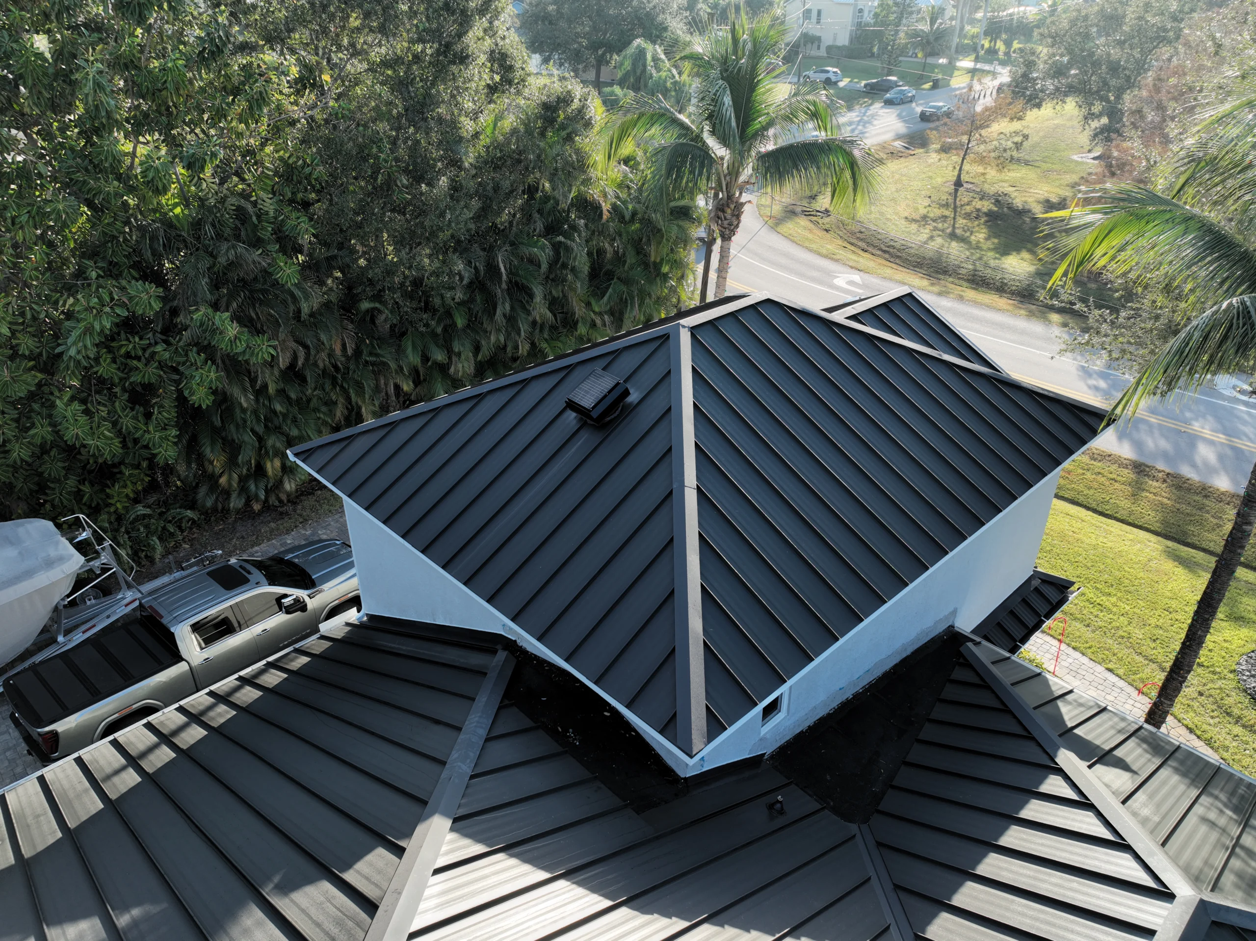 Aerial view of a house with a modern, black metal roof featuring multiple peaks and ridges. The house is surrounded by lush greenery and palm trees. A road with sparse traffic is visible in the background, along with a parked vehicle.