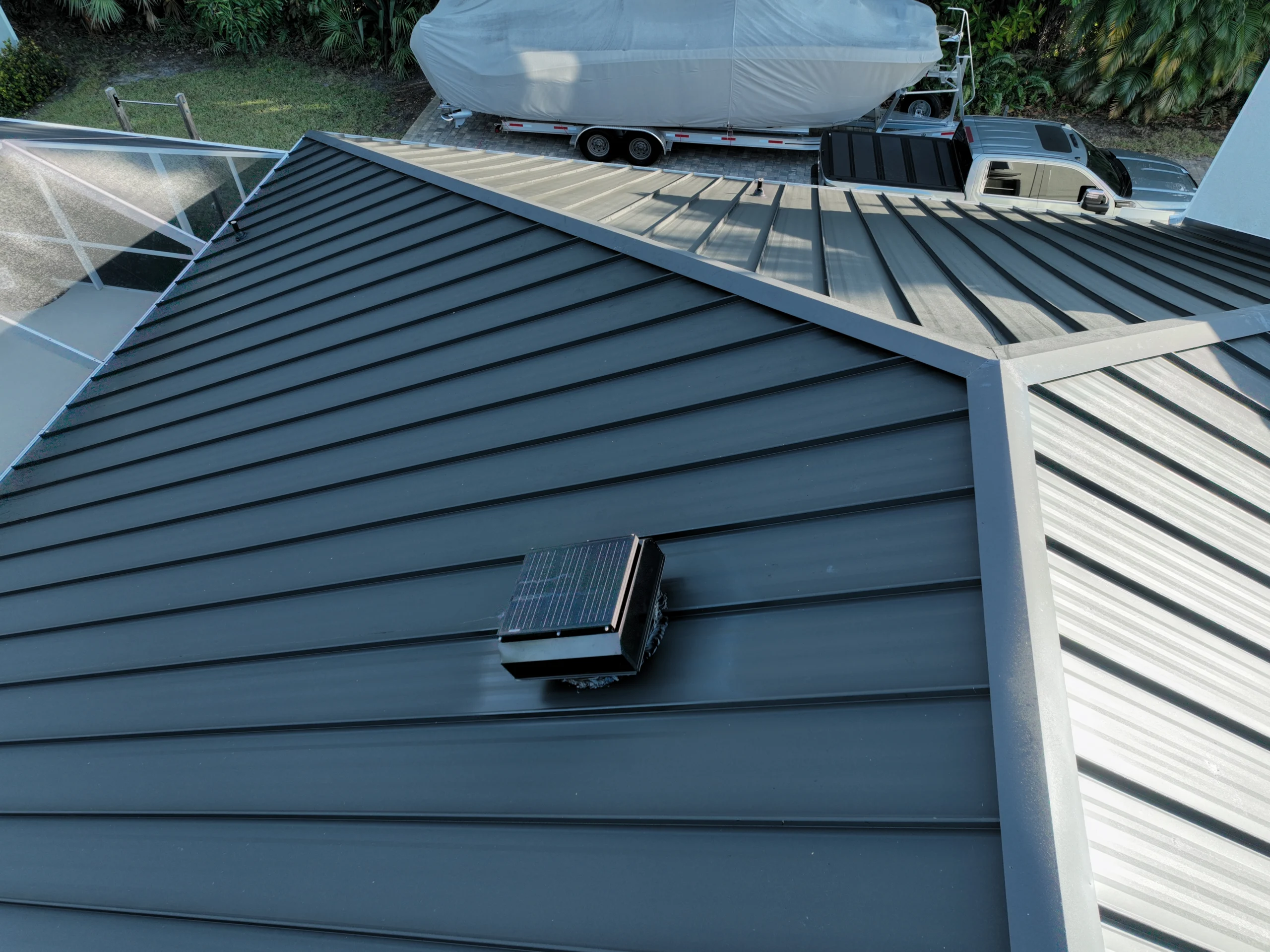 A metal roof with a small solar panel unit sits atop a building. In the background, there's a boat on a trailer and a white vehicle parked nearby, surrounded by greenery.