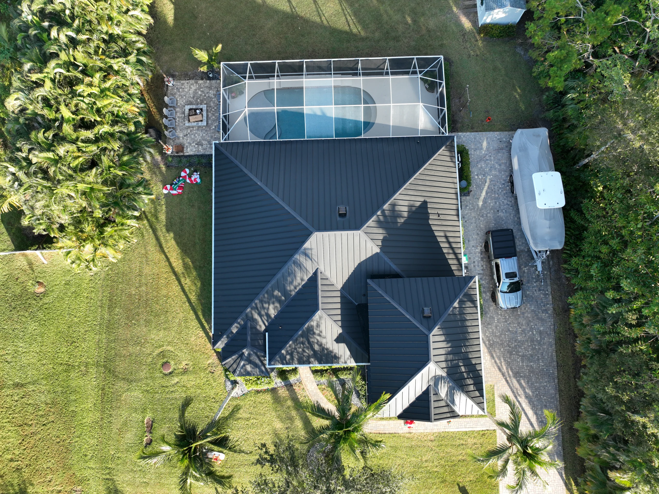 Aerial view of a modern house with a dark roof, surrounded by green lawns and palm trees. A screened-in pool is visible at the back. A driveway with a parked car and a recreational vehicle is on the right.