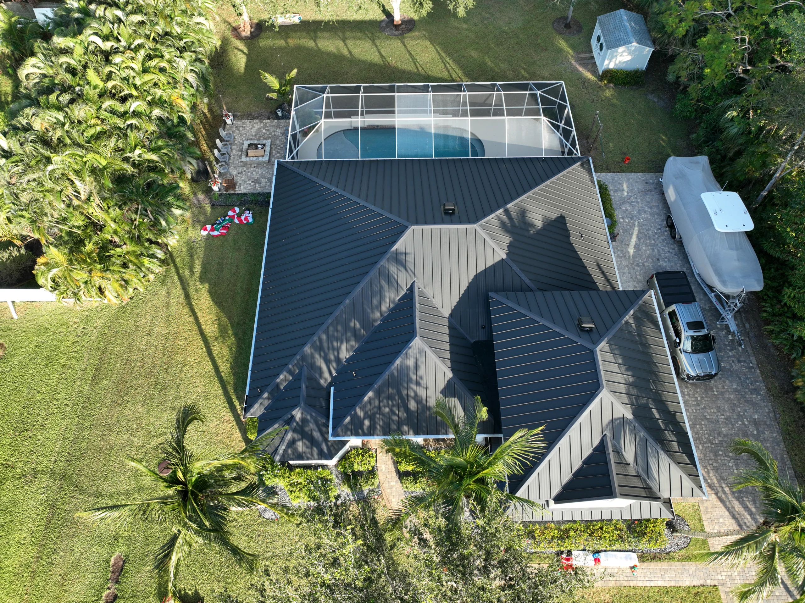 Aerial view of a house with a black metal roof and a screened pool area. The yard is surrounded by trees and plants. A parked vehicle and a trailer are visible on a paved driveway.