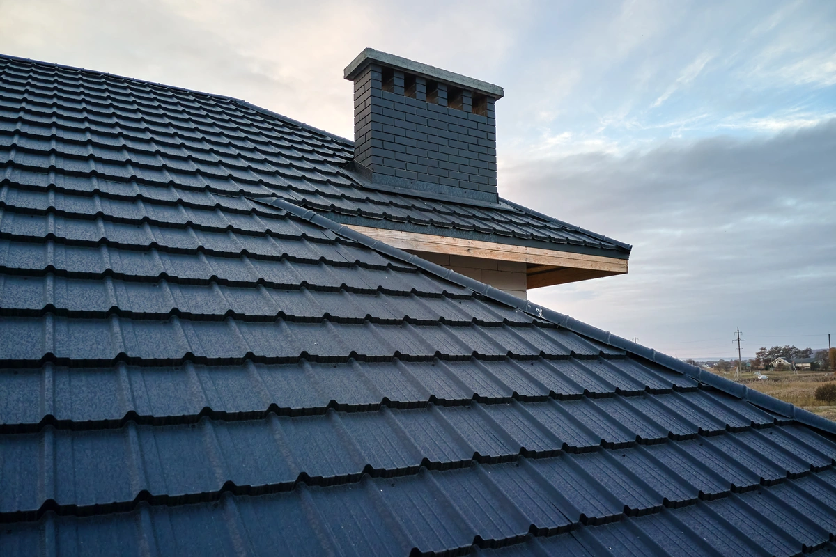 Close-up view of a roof with dark gray, textured metal tiles showcasing an innovative roof coating. A chimney in the same color scheme emerges from the roof. The sky is overcast with clouds, and glimpses of a distant landscape appear to the right.