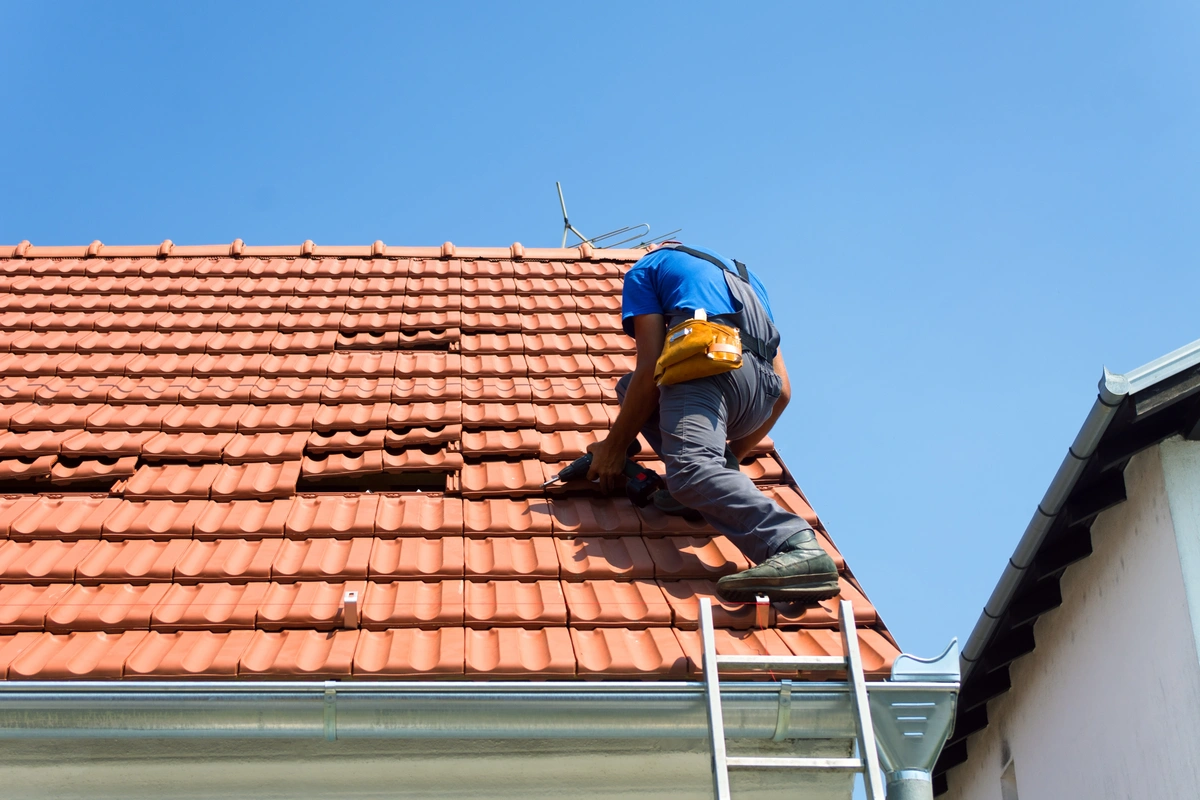 A person wearing a blue shirt and gray pants is busy with tile roof repair, standing on a ladder equipped with a tool belt. Several red tiles are missing against the backdrop of a clear, blue sky.