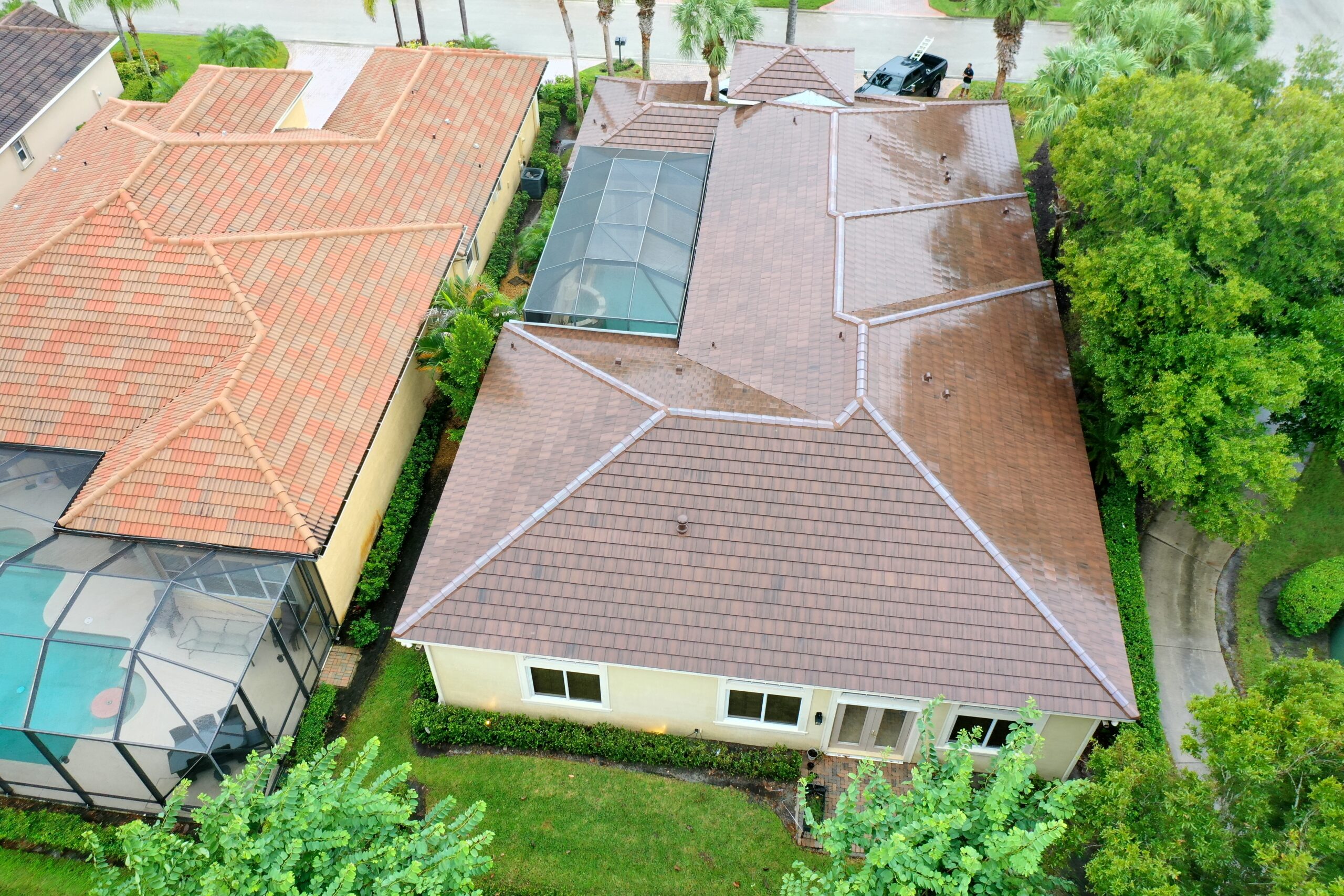 Aerial view of two suburban homes with upgraded brown tiled roofs, separated by greenery. One home boasts a screened pool enclosure. Wet pavement and lush trees suggest recent rain, with a black vehicle parked nearby.
