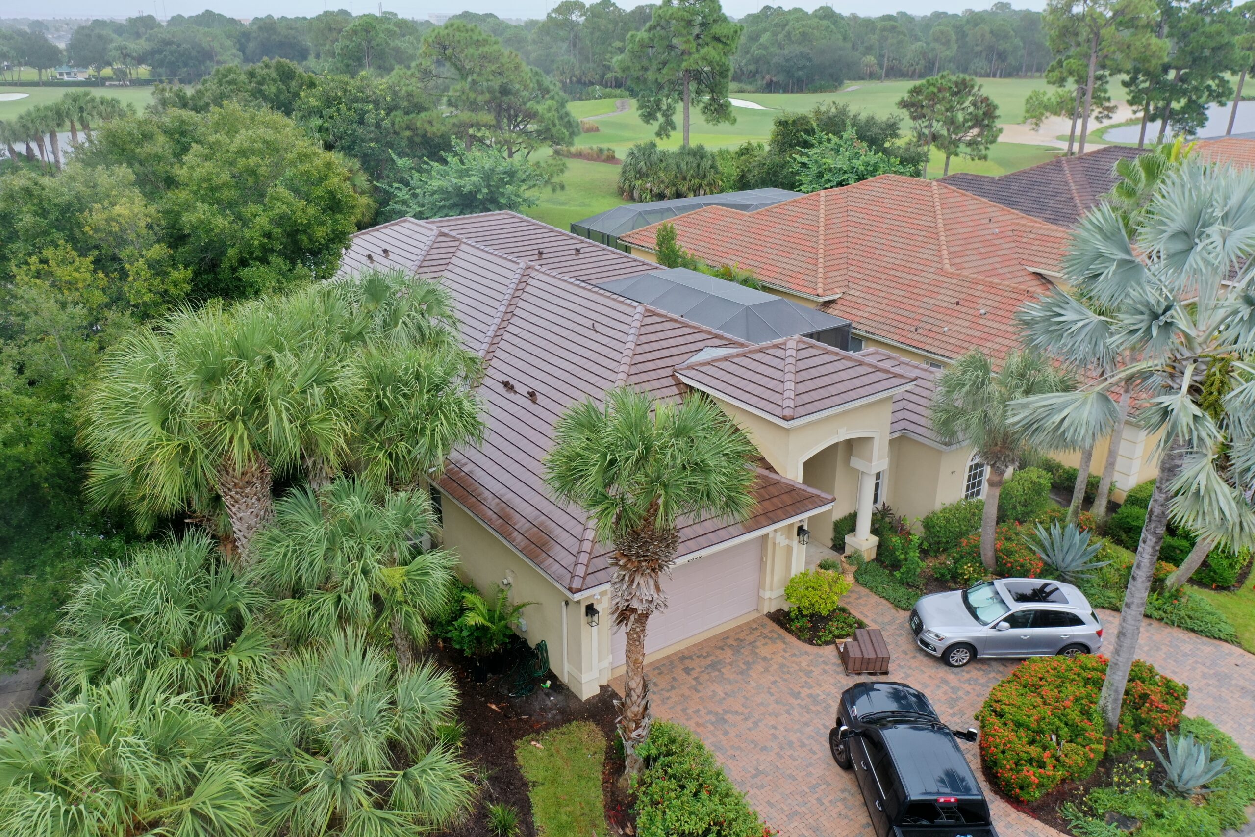 An aerial view reveals a house with a freshly upgraded tiled roof, nestled among lush palm trees and greenery. Two cars rest on the paved driveway. Nearby, similar houses line the surroundings, bordered by an expansive golf course dotted with sand traps.