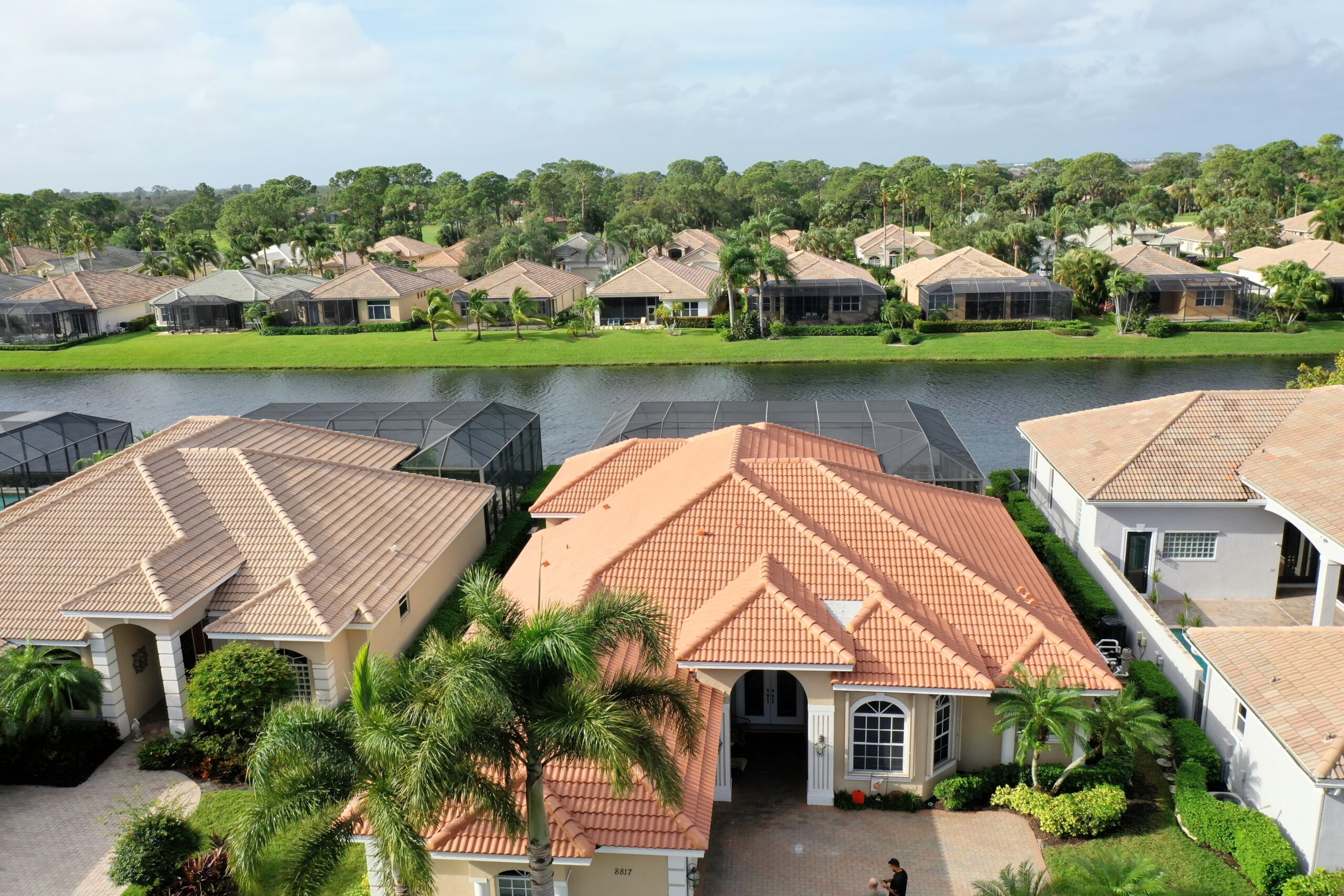 Aerial view of Caramel Estate in Port Saint Lucie, showcasing suburban homes with concrete tile roofs. A canal divides the neatly arranged houses, framed by lush palm trees and vibrant greenery under an overcast sky.