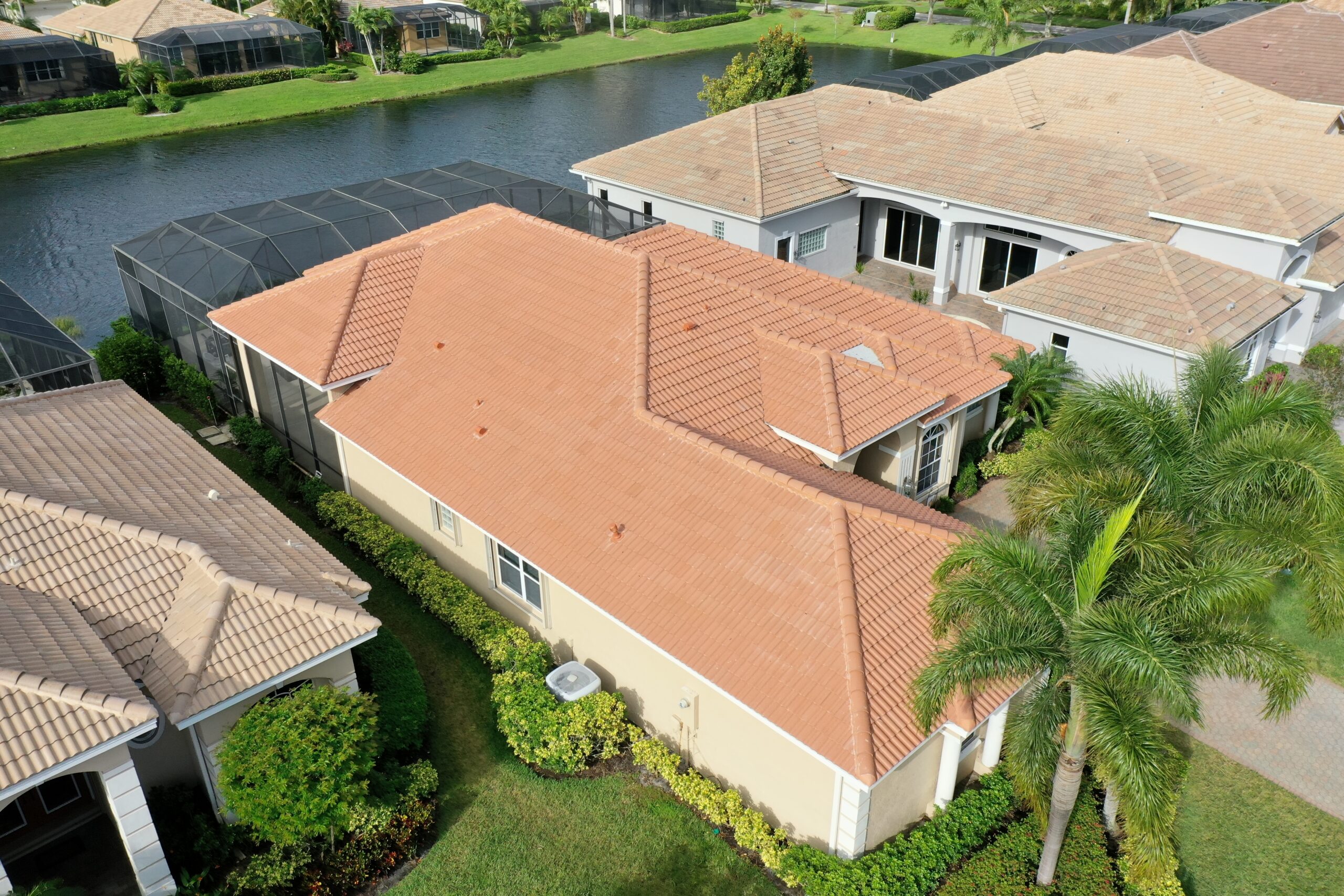Aerial view of the picturesque Caramel Estate neighborhood in Port Saint Lucie, showcasing a tan house with a terracotta roof surrounded by manicured lawns. A tranquil canal bordered by other charming homes with concrete tile roofs is visible in the background.