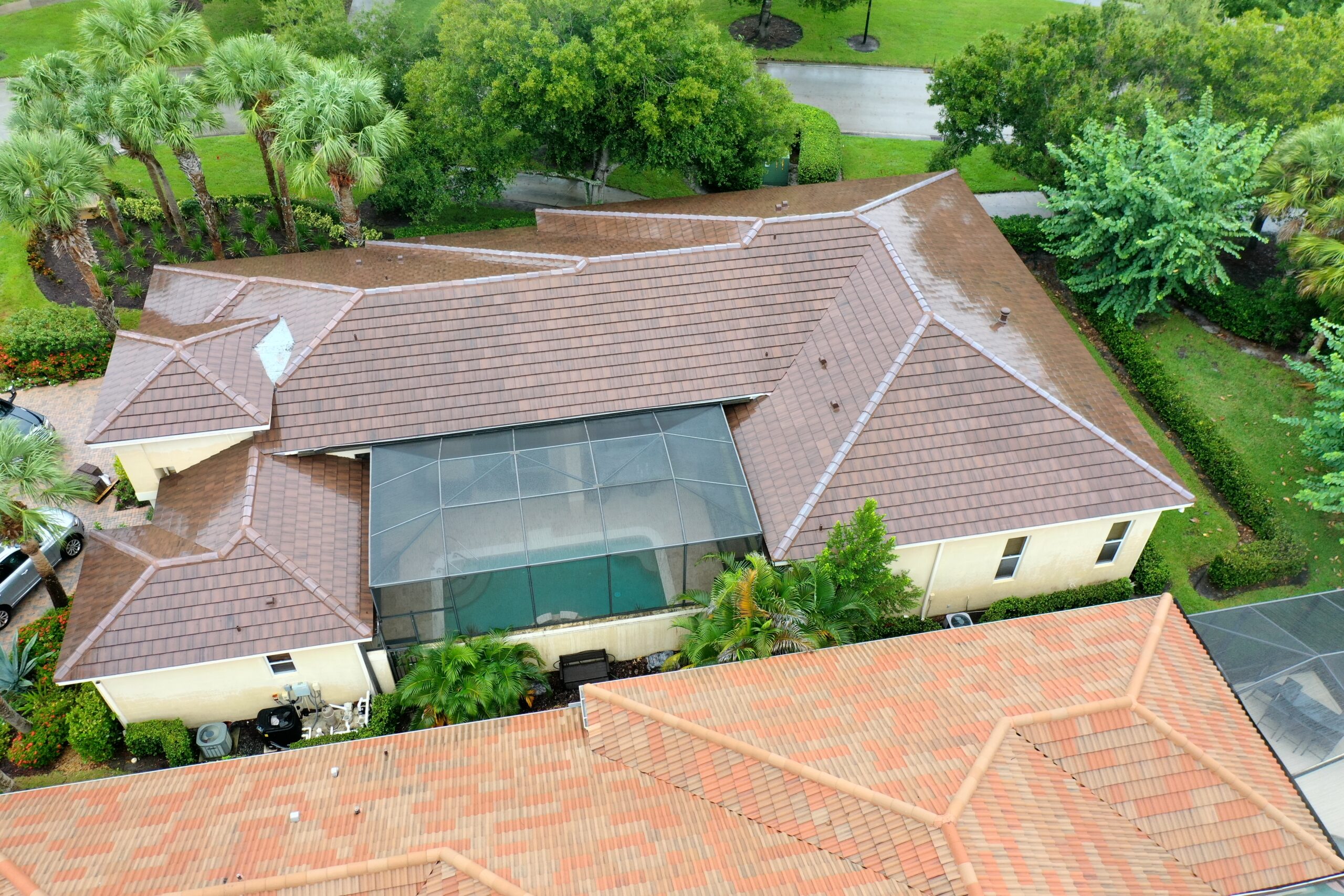 Aerial view of a house with a brown tile roof, showcasing its recent roof upgrade. The central feature is a screened-in pool area. Green trees and a driveway surround the house, while another roof is partially visible in the foreground.