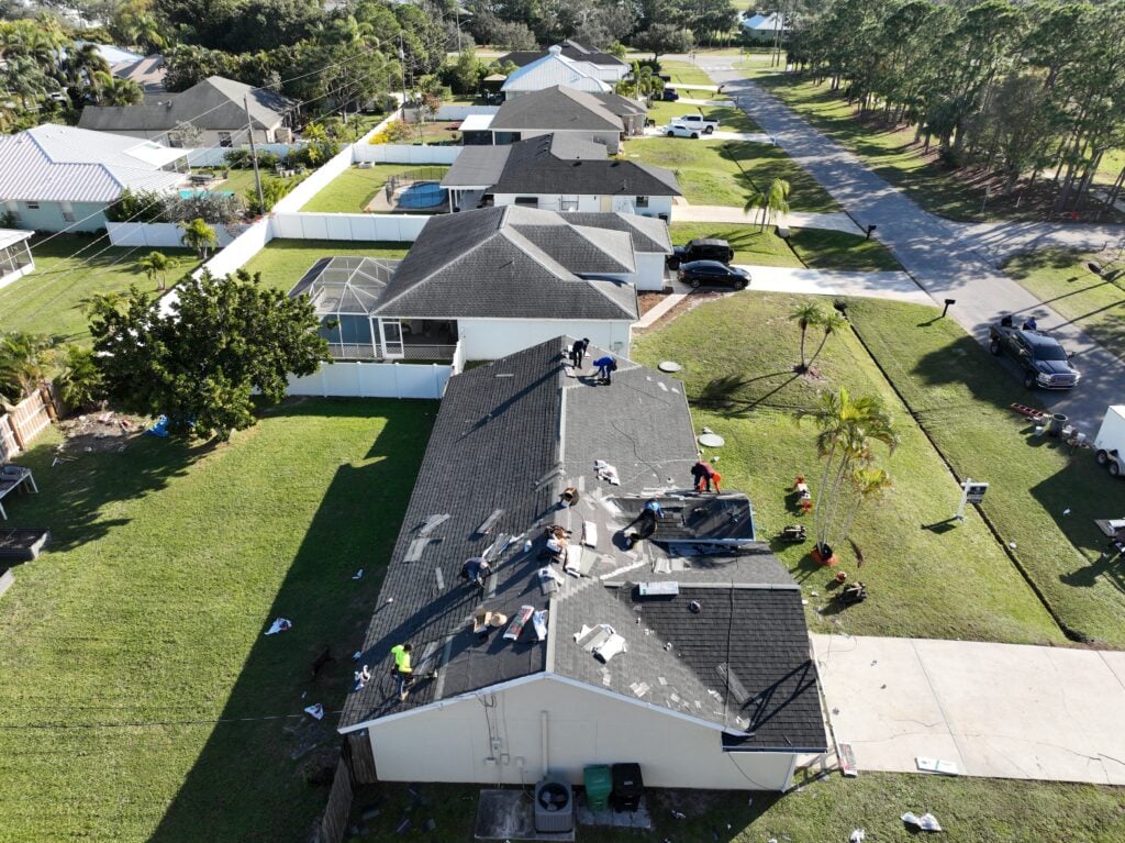 Aerial view of workers providing Shingle Roofing Services, repairing the roof of a house in a suburban treasure coast neighborhood. Tools and materials are scattered around, with similar homes and green lawns lining the street in the background.