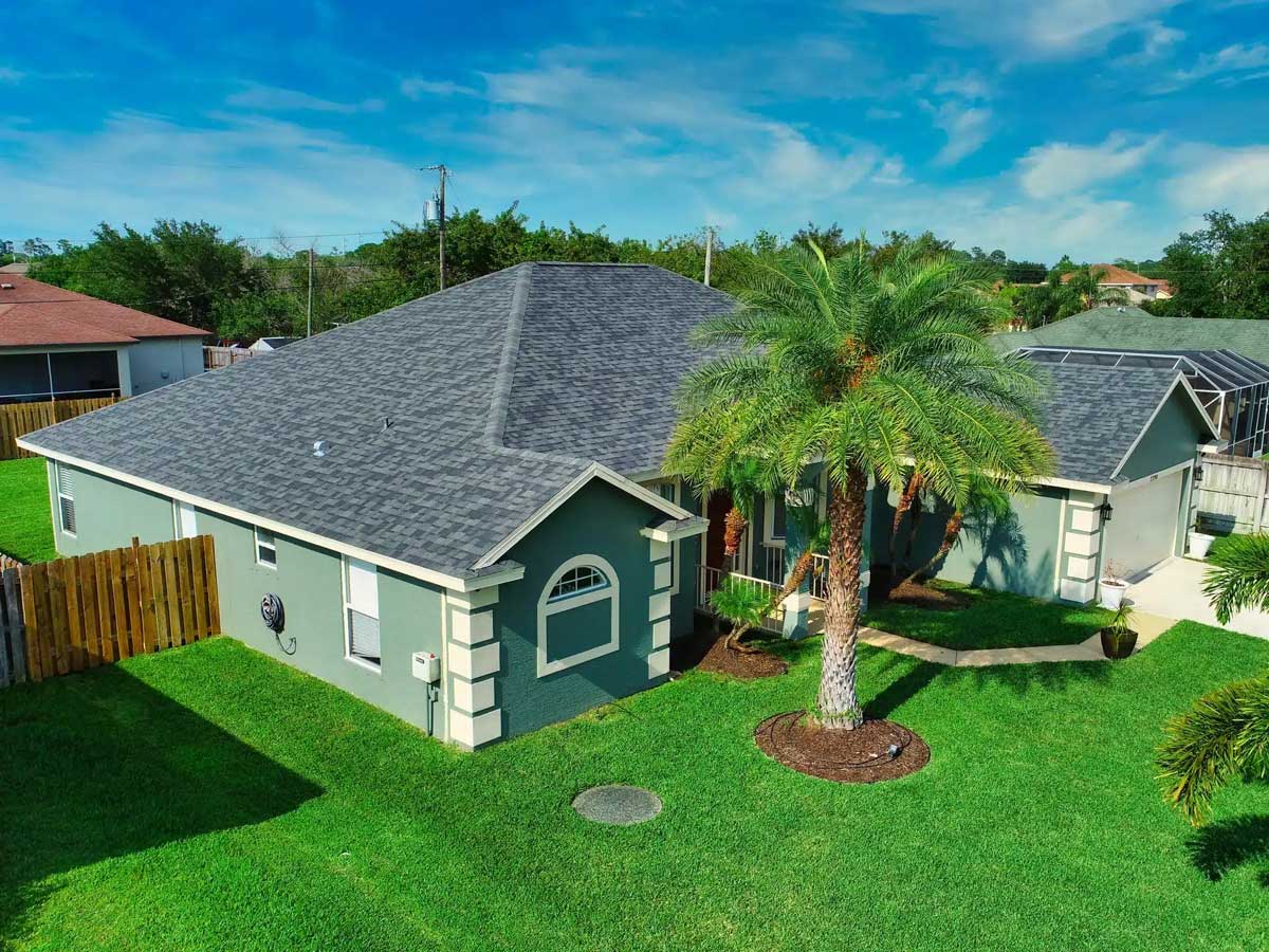 A single-story green house with a gray shingle Rhino Roofs system, arched window, attached garage, and a manicured lawn with palm trees, set under a bright blue sky. Ideal for quality residential roofing.