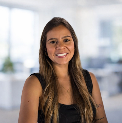 A woman with long brown hair and a black sleeveless top smiles while sitting in a bright, modern office with blurred desks and windows in the background.