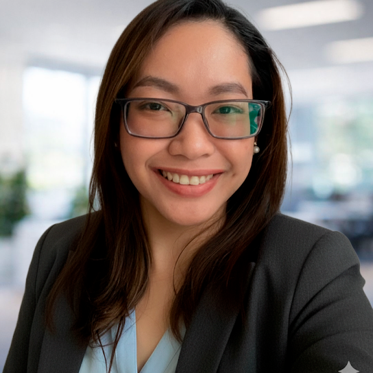 A woman with long brown hair and glasses, wearing a blazer and smiling, stands in a brightly lit office with large windows in the background.