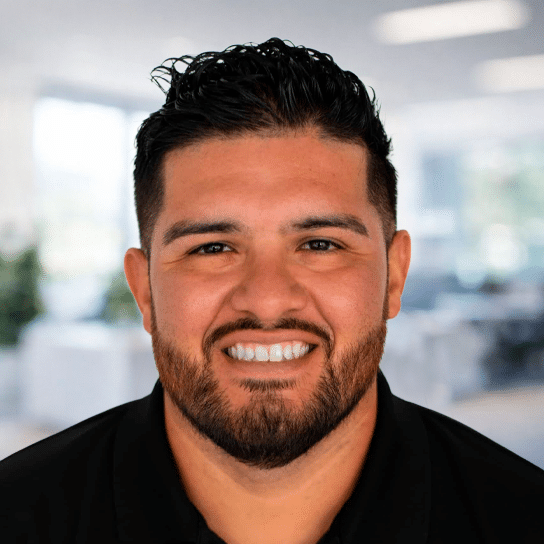 A man with short dark hair and a beard smiles at the camera. He is wearing a black collared shirt and is standing in a bright, modern office setting.