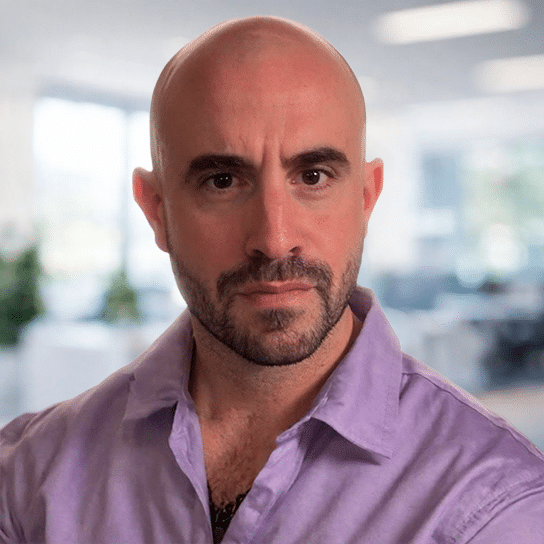 A bald man with a trimmed beard wearing a lavender collared shirt stands indoors, looking directly at the camera with a serious expression. The blurred background suggests an office environment.