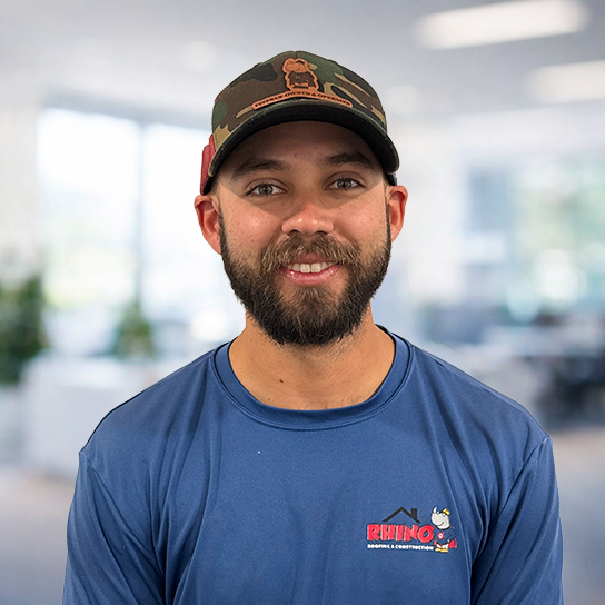 A man with a beard smiles at the camera. He wears a camouflage cap with a dog patch and a blue 