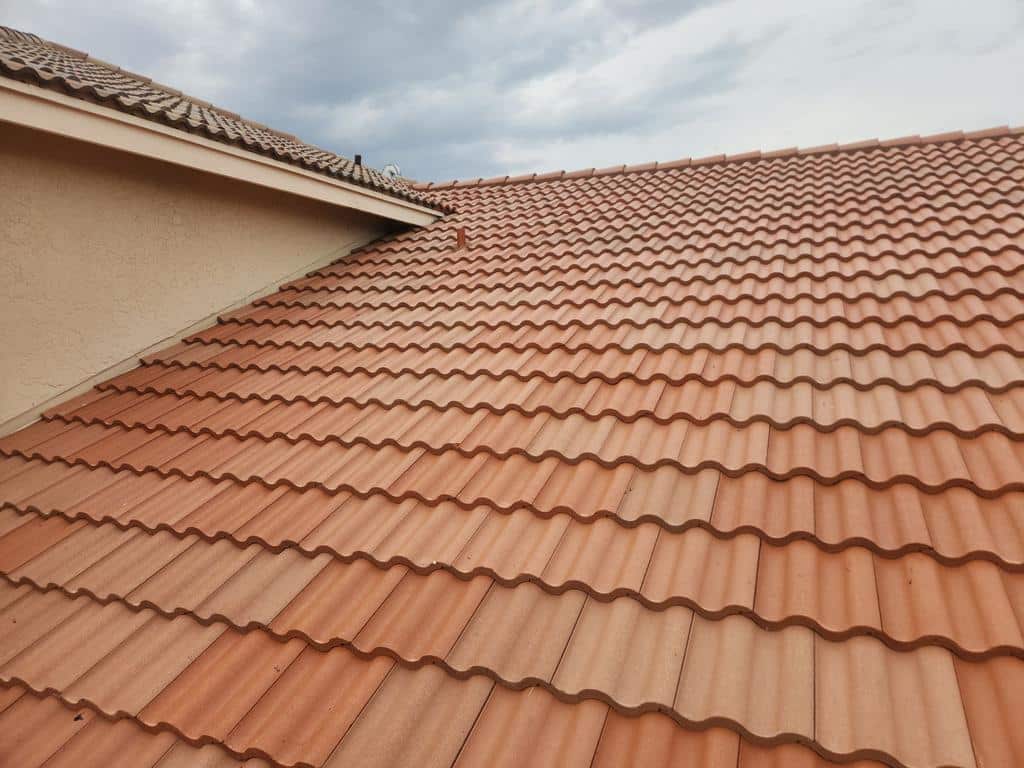 A close-up view of a house roof with red-orange curved clay tiles under a cloudy sky.
