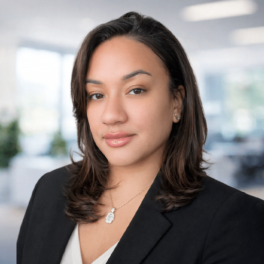 A woman with shoulder-length brown hair wearing a black blazer, white blouse, and a gold necklace stands in an office setting with a blurred background.