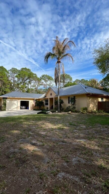 A single-story yellow house with a gray roof and garage, a tall palm tree in the front yard, and a backdrop of pine trees under a blue sky with scattered clouds.