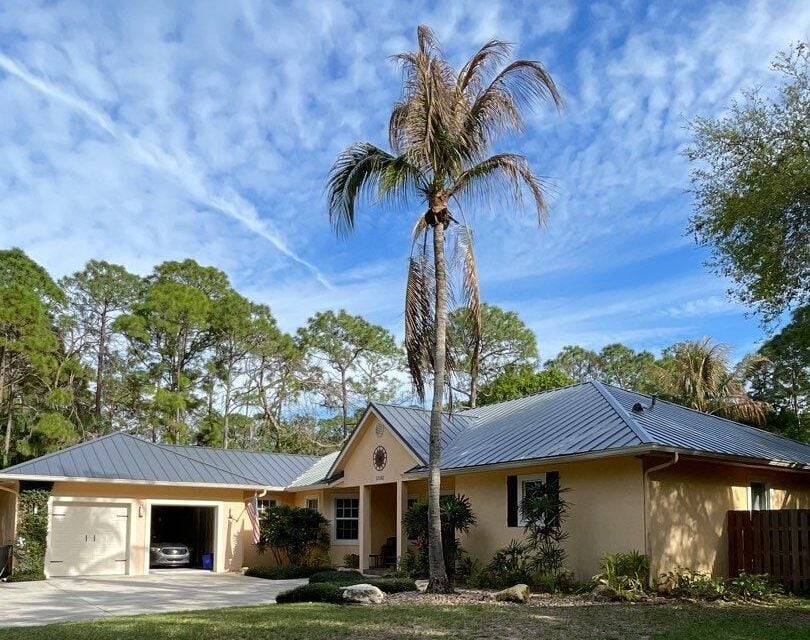 A single-story yellow house with a gray roof and garage, a tall palm tree in the front yard, and a backdrop of pine trees under a blue sky with scattered clouds.