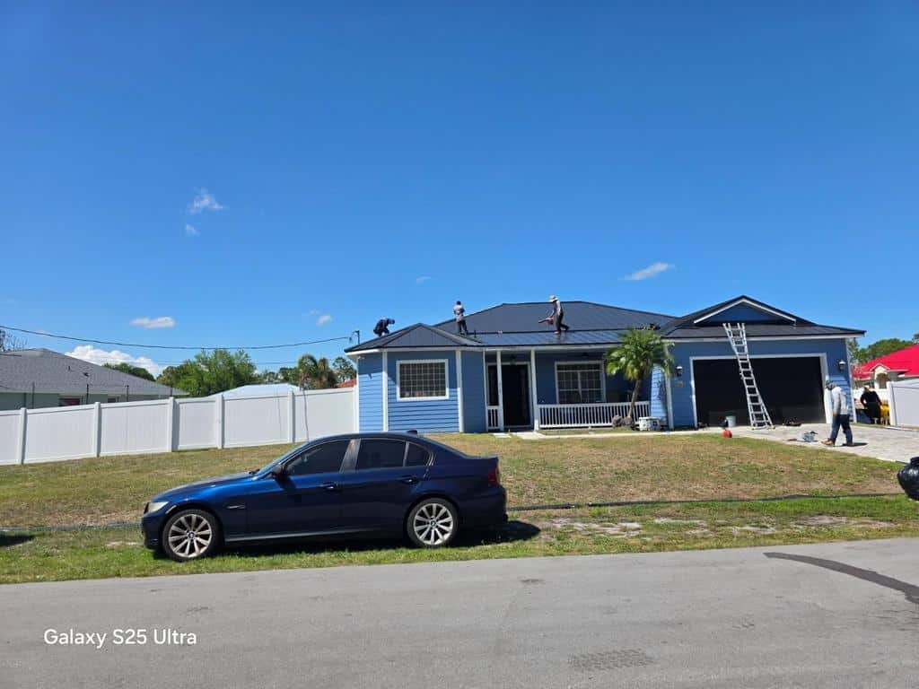 A blue car is parked in front of a blue house with three people working on its roof. A ladder leans against the garage, and the sky is clear and sunny. The words “Galaxy S25 Ultra” appear in the lower left.