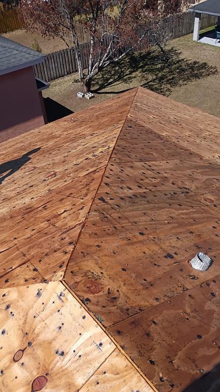 A rooftop covered with plywood sheets, showing the wood grain and seams. There is a metal vent on one side and a yard with trees and a fence in the background.