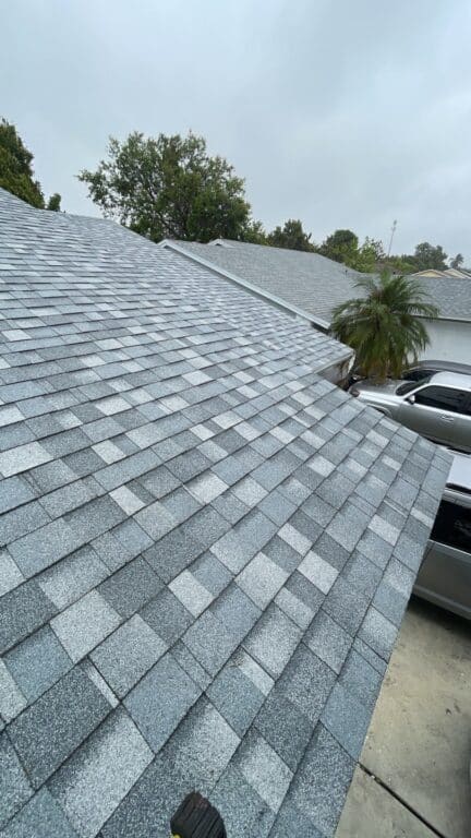 A close-up view of a gray shingled roof on a cloudy day, with trees, a palm tree, and parked cars visible in the background.