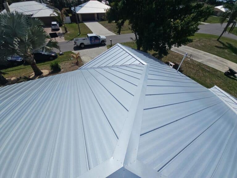Aerial view of a shiny silver metal roof with distinct seams and panels; residential neighborhood with trees, houses, and a pickup truck visible below.