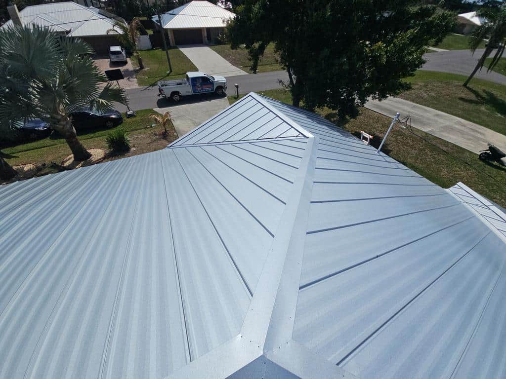Aerial view of a shiny silver metal roof with distinct seams and panels; residential neighborhood with trees, houses, and a pickup truck visible below.