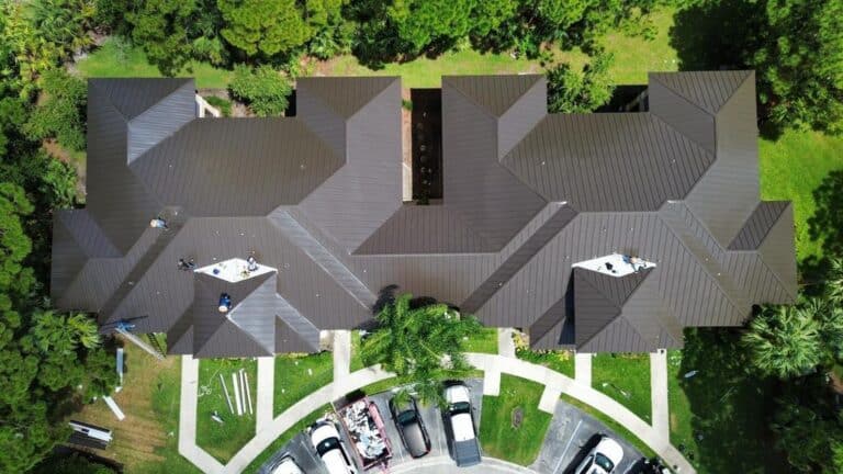 Aerial view of a large building with a dark metal roof, several workers present. Trees surround the structure, and a parking lot with cars is visible at the bottom of the image.