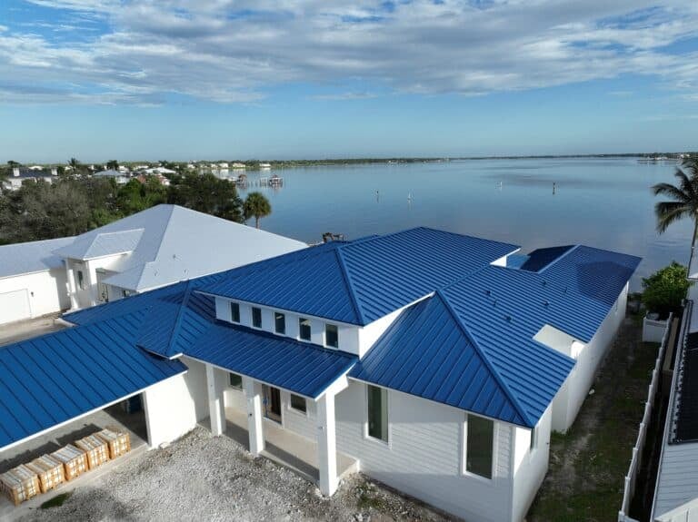 Modern white house with a blue metal roof sits beside calm water under a partly cloudy sky. Nearby, more houses and trees line the waterfront, with a palm tree on the right and construction materials in the foreground.